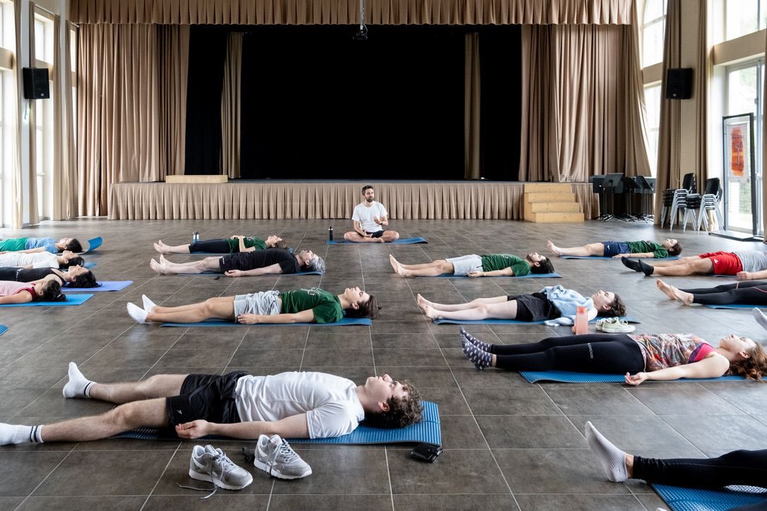 in a large hall with a stage, alex sits cross-legged on a mat while about fifteen people lie on their backs on blue mats around him, relaxing.